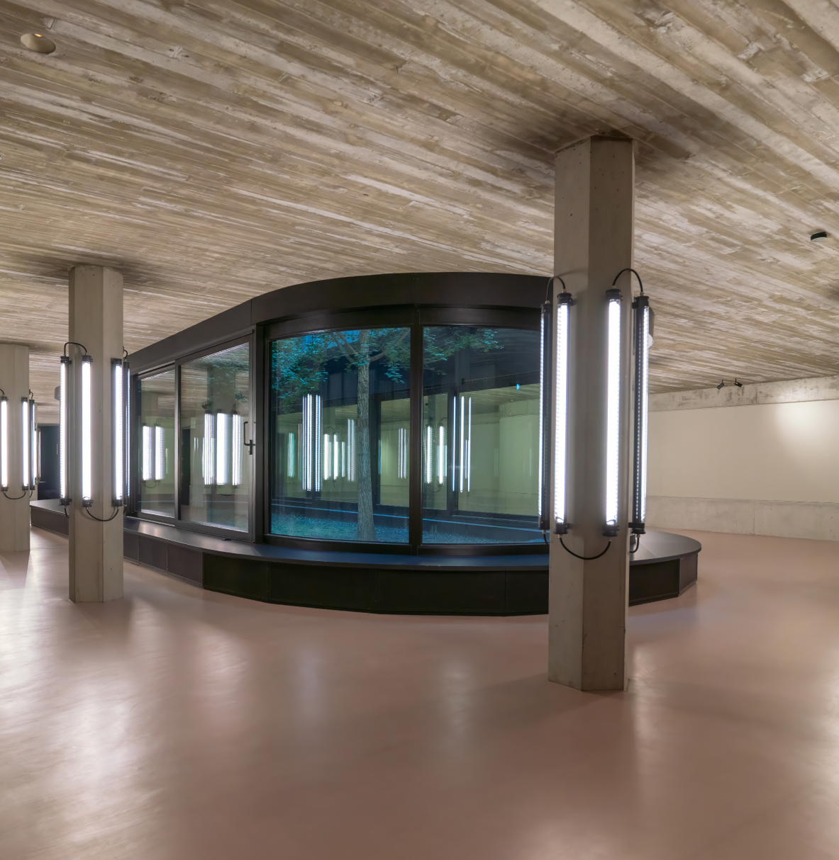 Moderner Innenraum mit Betonwänden und -decken, einem runden Glaskasten in der Mitte mit Bäumen darin und beleuchteten Säulen mit vertikalen Lichtstäben.