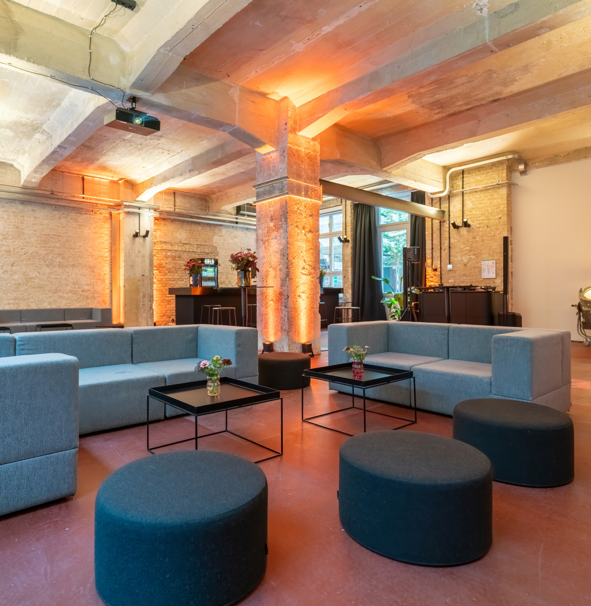 Moderner Lounge-Bereich mit zwei blauen Sofas, schwarzen quadratischen Couchtischen mit kleinen Blumengestecken und mehreren runden, dunklen Poufs in einem Loft mit Sichtbetondecke und Ziegelwänden, akzentuiert durch warme orange Beleuchtung an den Säulen und Wänden.