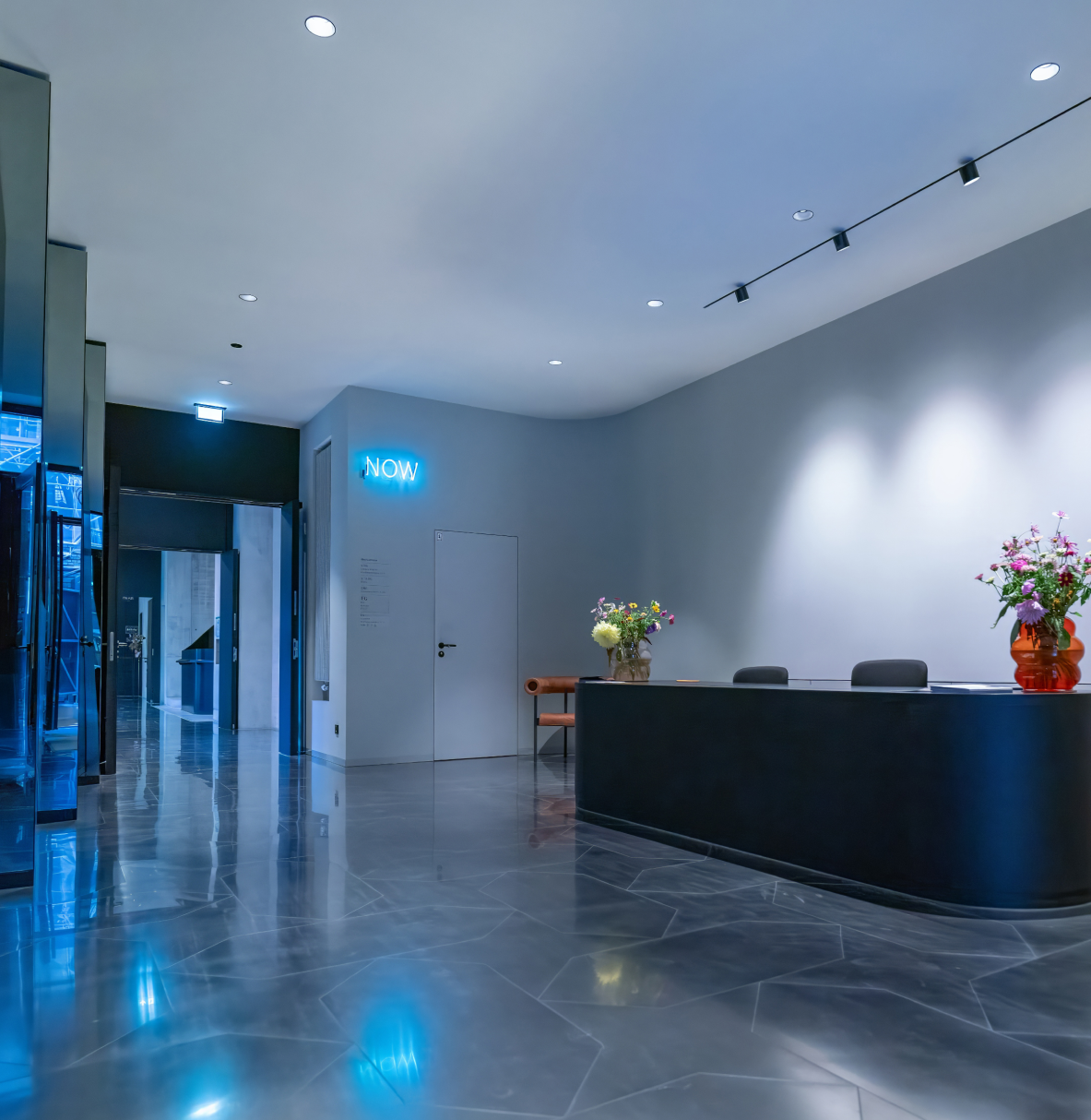 Moderner Empfangsbereich mit glänzendem Marmorfußboden, schwarzem Empfangstresen mit zwei Vasen bunter Blumen und einem orangenen Stuhl an grauer Wand mit blauem Neonzeichen "NOW".
