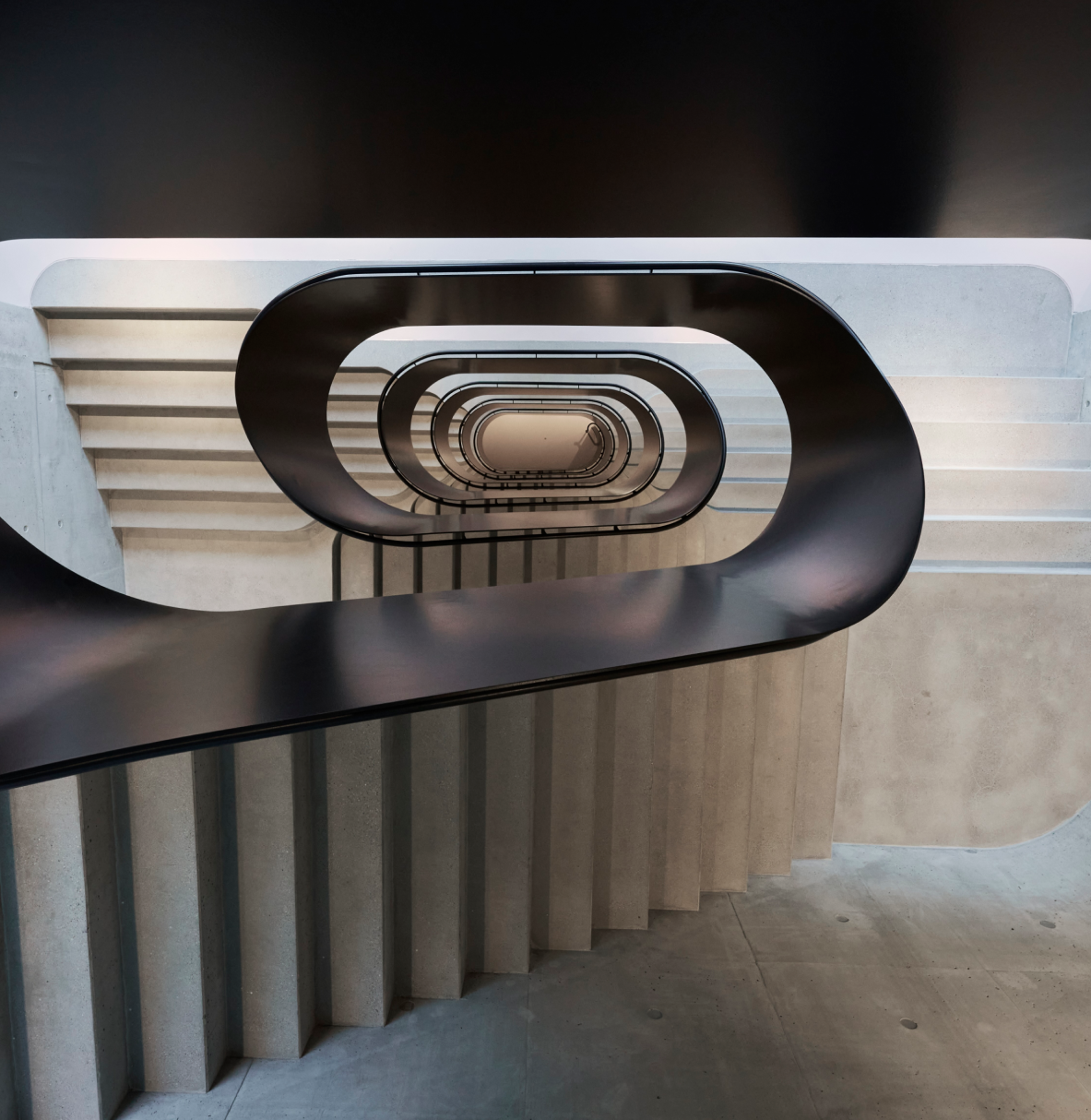 Blick von oben auf eine moderne Wendeltreppe mit schlichtem, schwarzen Geländer und grauen Betonstufen in einem Gebäude.