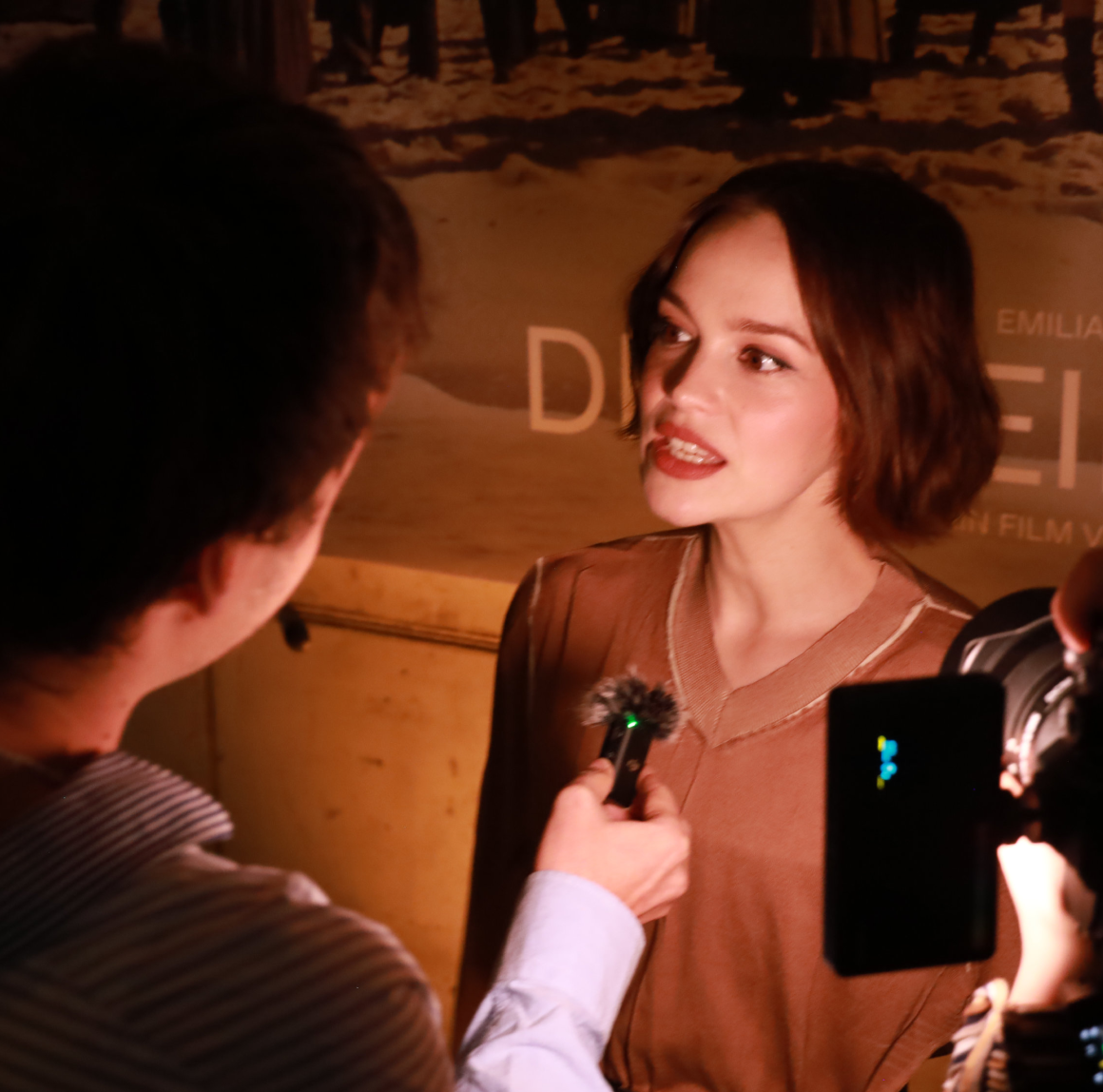 Eine Frau mit braunen, schulterlangen Haaren in einem braunen Oberteil spricht während eines Interviews, während eine andere Person vor ihr ein Mikrofon hält. Rechts ist eine Kamera mit Lichtquelle zu sehen.