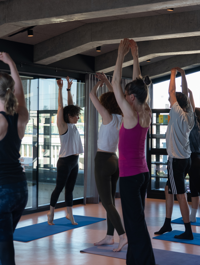 Eine Gruppe von Männern und Frauen, die gemeinsam in einem modernen Raum auf Yogamatten stehen und sich mit ausgestreckten Armen nach oben strecken, während sie Yoga oder Dehnübungen machen.