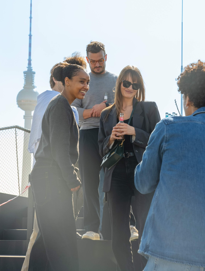 Fünf junge Erwachsene unterhalten sich und lachen auf einer Außentreppe an einem sonnigen Tag vor dem Berliner Fernsehturm. Zwei Frauen stehen vorn, eine mit Sonnenbrille hält eine Flasche, andere lächelt. Ein Mann und zwei weitere Personen sind im Hintergrund mit Getränken.
