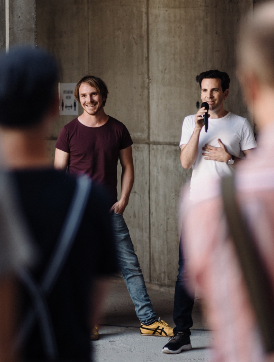 Zwei Männer stehen vor einer Betonwand; der eine trägt ein weißes T-Shirt und spricht in ein Mikrofon, der andere trägt ein dunkelrotes T-Shirt und lächelt, während zwei unscharfe Figuren im Vordergrund zuhören.