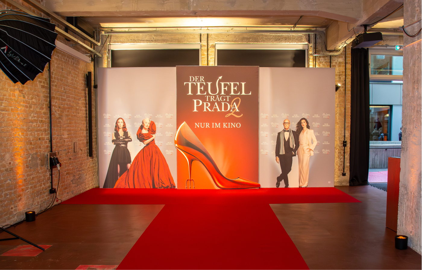 Roter Teppich vor einer Fotowand mit großem Plakat für den Film „Der Teufel trägt Prada 2“ mit einer roten High-Heel-Schuhskulptur und abgebildeten Schauspielern.