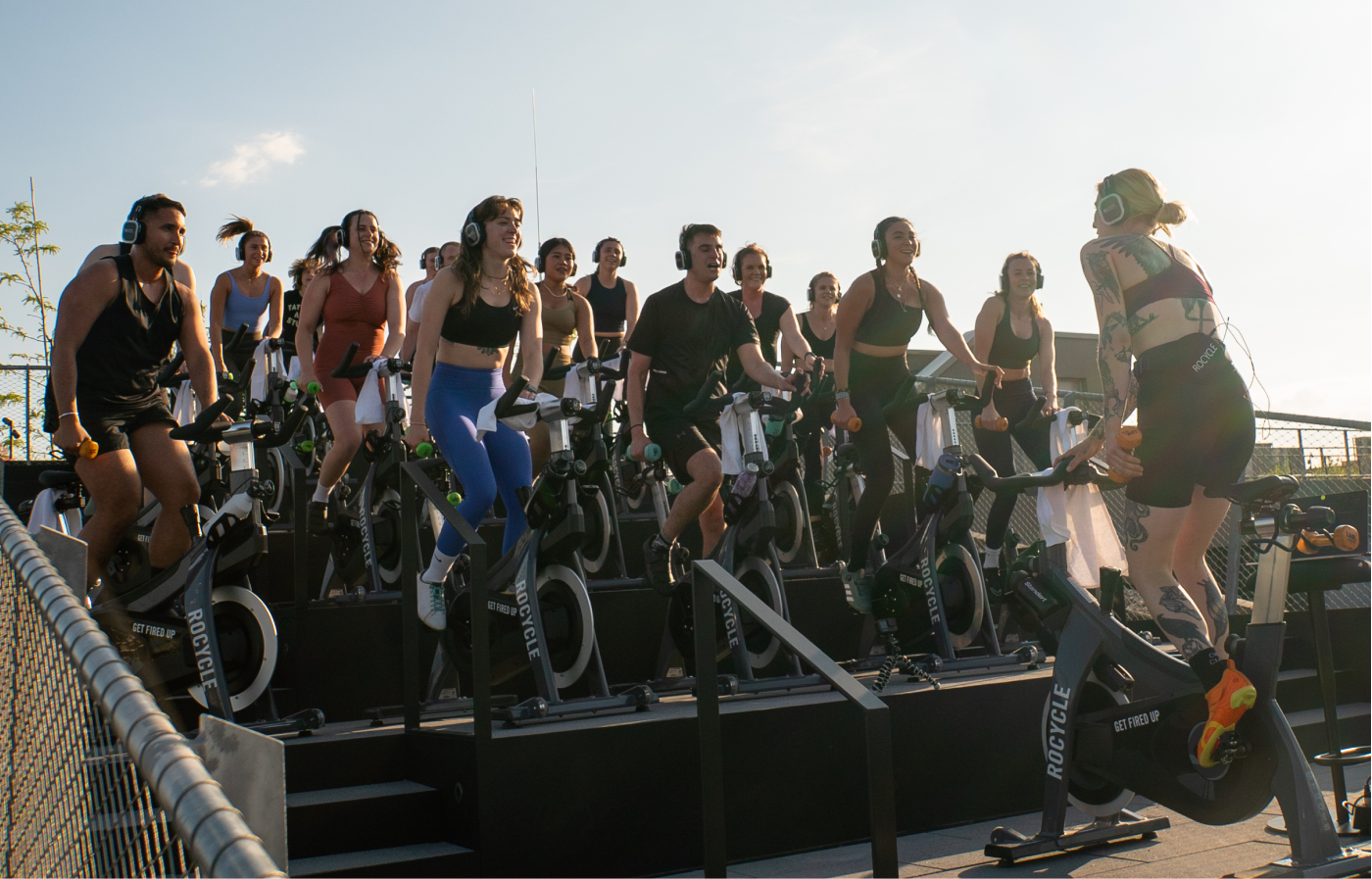 Gruppe von Menschen, die in einem Outdoor-Fahrradkurs auf Spinning-Bikes mit Kopfhörern trainieren, angeleitet von einer Trainerin mit Tattoos und orangen Sportschuhen.