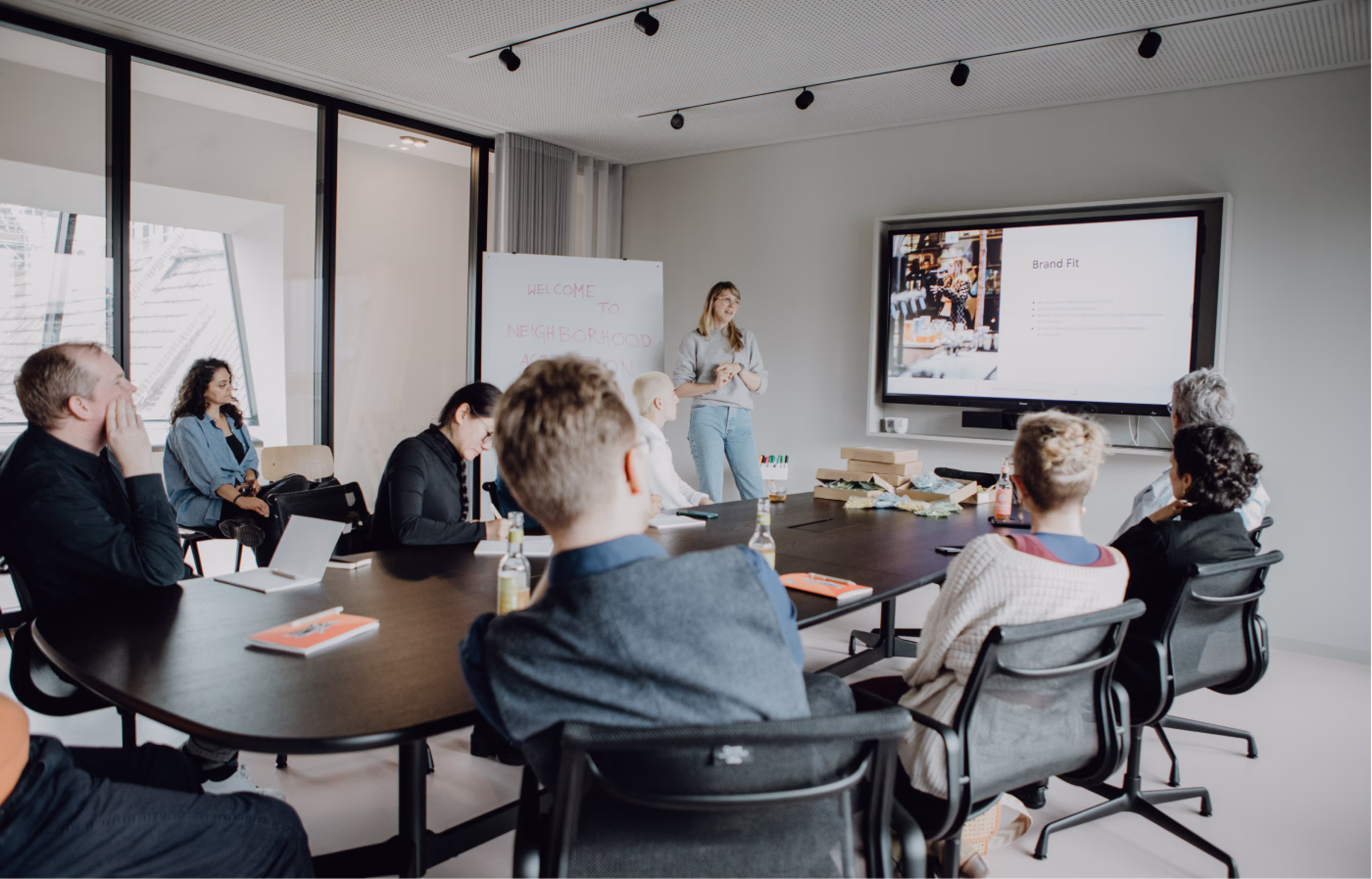 Eine Gruppe von acht Personen sitzt um einen Konferenztisch und hört einer Frau zu, die vorne stehend eine Präsentation mit einer großen Bildschirmanzeige hält.