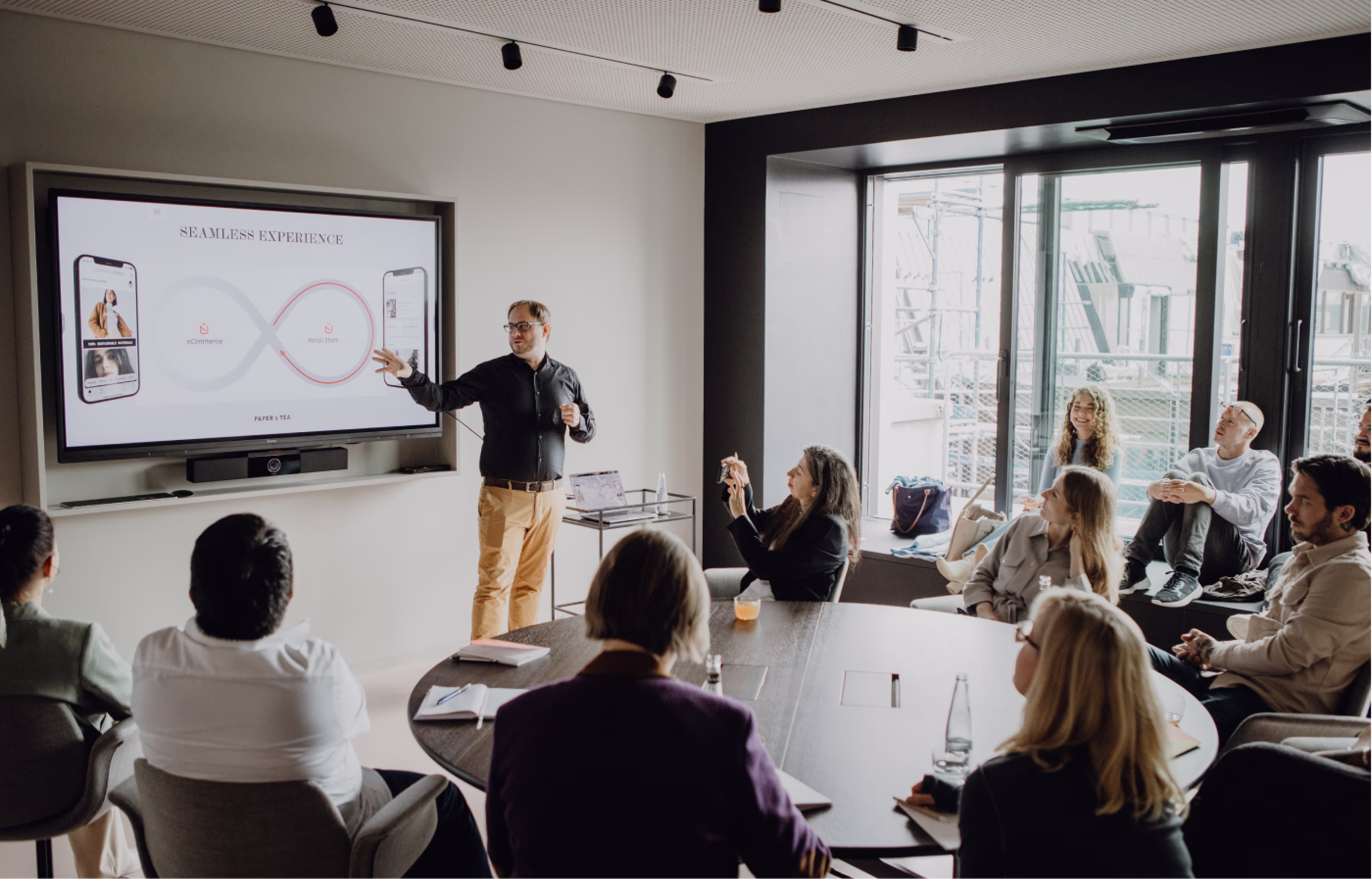 Ein Mann hält eine Präsentation in einem Konferenzraum vor mehreren Personen, die an einem Tisch sitzen oder auf einer Fensterbank. Auf dem Bildschirm ist eine Grafik mit der Überschrift 'SEAMLESS EXPERIENCE' und zwei Smartphone-Darstellungen zu sehen.
