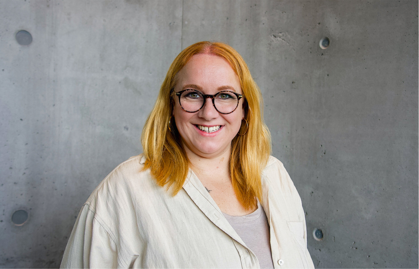 Frau mit roten Haaren, runder Brille und hellem, lockeren Hemd lächelt vor einer grauen Betonwand.