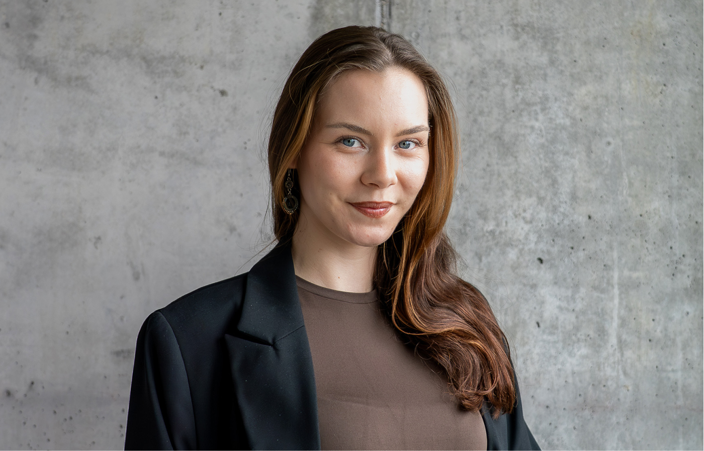 Porträt einer lächelnden jungen Frau mit langen braunen Haaren, die einen schwarzen Blazer und ein braunes Oberteil trägt, vor einer grauen Betonwand.