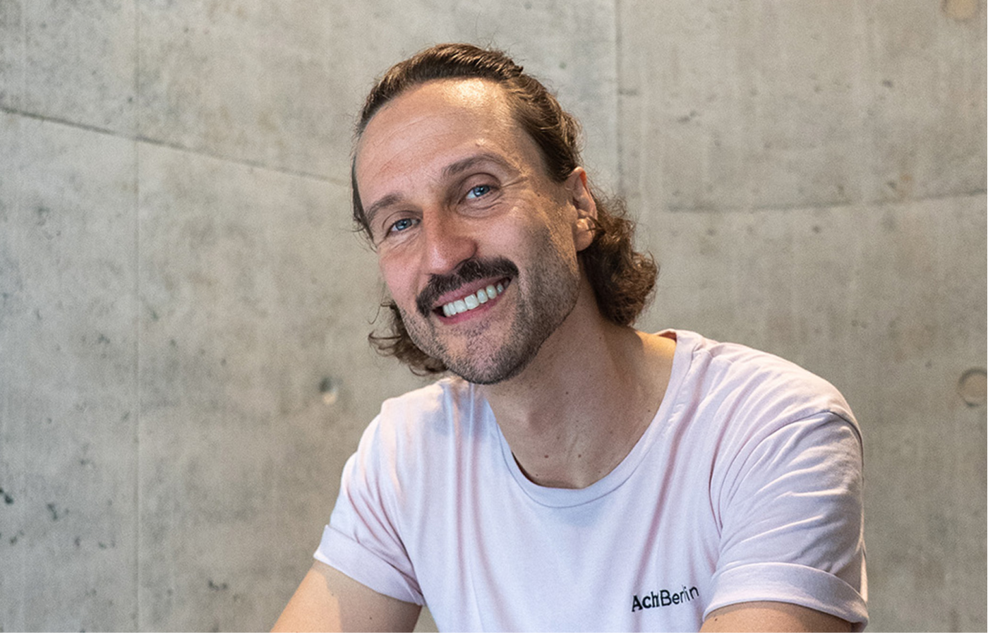 Lächelnder Mann mit Schnurrbart und welligem, schulterlangem Haar trägt ein helles T-Shirt mit der Aufschrift „AchtBerlin“ vor einer grauen Betonwand.