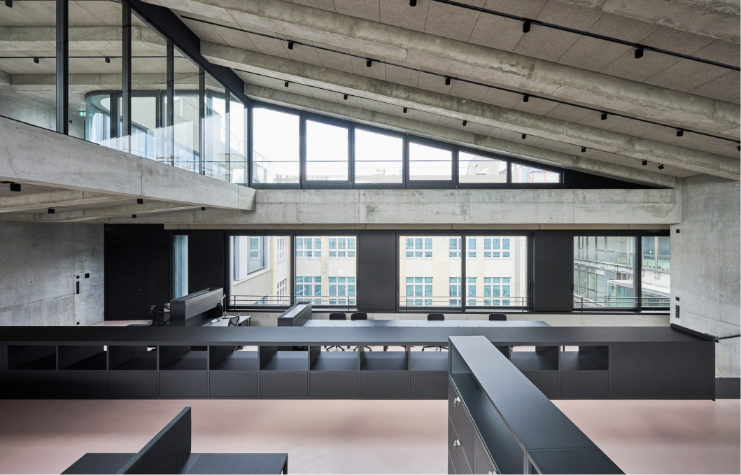 Modernes Büro mit sichtbaren Betonwänden, großen Fenstern und einem geneigten Dachtragwerk aus Beton. Innen stehen schwarze Möbel und Schreibtische mit Stühlen vor den Fenstern, darunter Regale und Schränke, die den Raum in Zonen aufteilen. Das Tageslicht fällt durch die Fenster und betont die industrielle, minimalistische Gestaltung des Raums.
