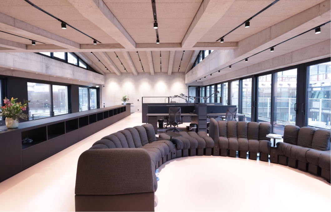 Modernes, offenes Büro mit großen Fenstern, die viel natürliches Licht hereinlassen, einer geschwungenen grauen Couch im Vordergrund und mehreren Arbeitsplätzen mit schwarzen Stühlen und Lampen im Hintergrund.