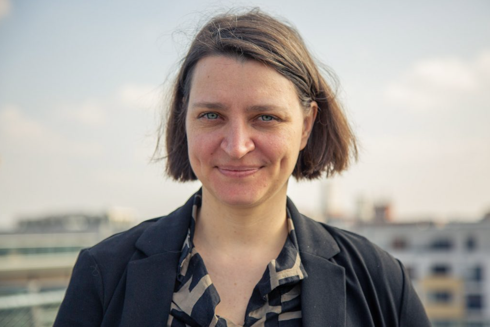 Portrait einer Frau mit kurzen braunen Haaren, die in die Kamera lächelt, vor einem unscharfen städtischen Hintergrund bei Tageslicht.