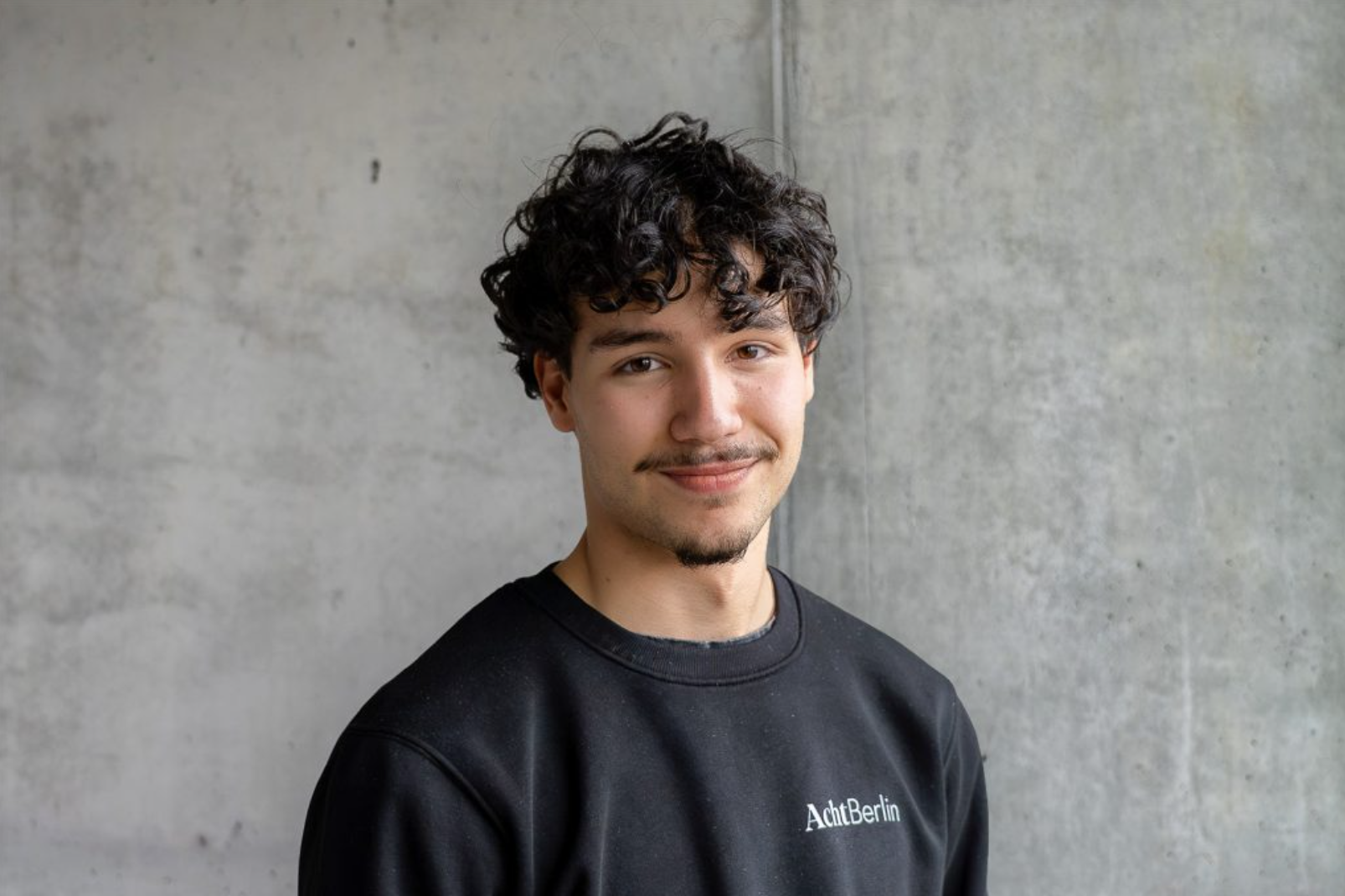 Junger Mann mit lockigem dunklem Haar, Schnurrbart und Kinnbart lächelt in die Kamera, trägt schwarzen Pullover mit Aufschrift "AchtBerlin" vor einer grauen Betonwand.