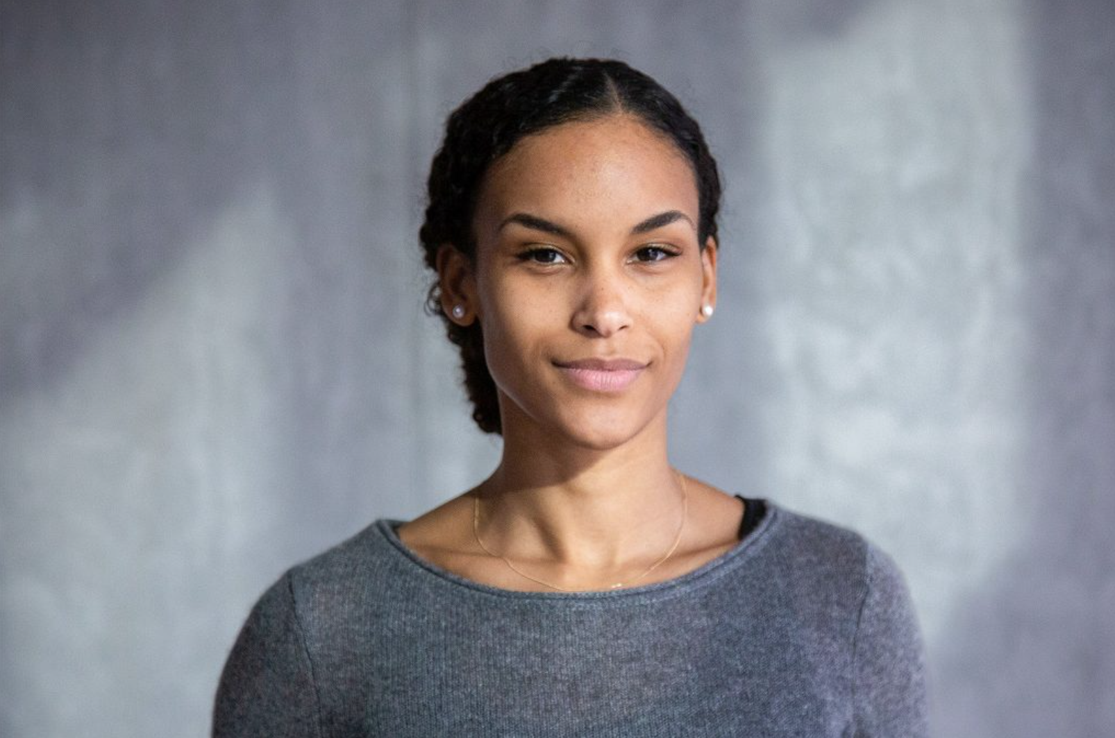 Junge Frau mit zurückgebundenem lockigem Haar, die in einem grauen Pullover vor einem grauen, unscharfen Hintergrund in die Kamera lächelt.