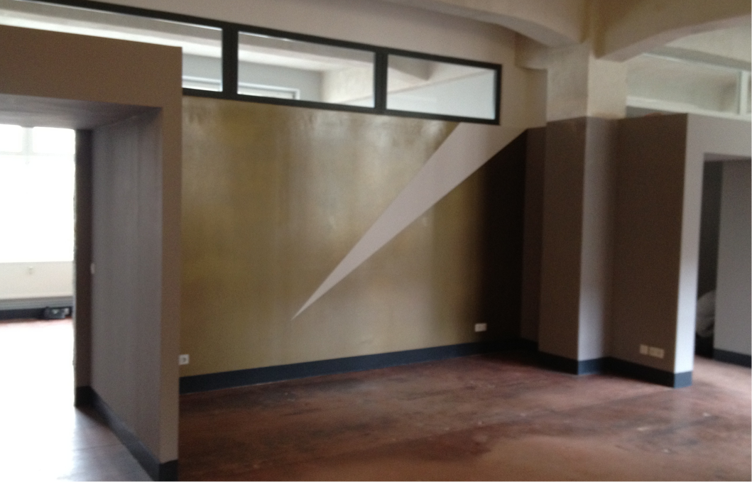 Leerer Raum mit braunem Boden und Wänden, die teilweise in Grau und Braun gestrichen sind. Eine Wand zeigt ein diagonales, hellgraues Dreieck als Wandgestaltung. Oben an der Wand sind Fenster, die in einen angrenzenden Raum mit mehr Tageslicht blicken lassen.