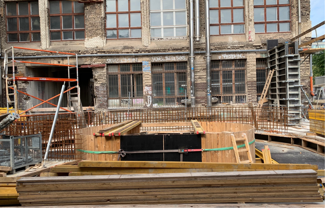 Baustelle vor einem alten, grauen Backsteingebäude mit großen Fenstern und Graffiti; im Vordergrund sind Holzstapel, Metallgitter und eine runde Holzverschalung sichtbar.