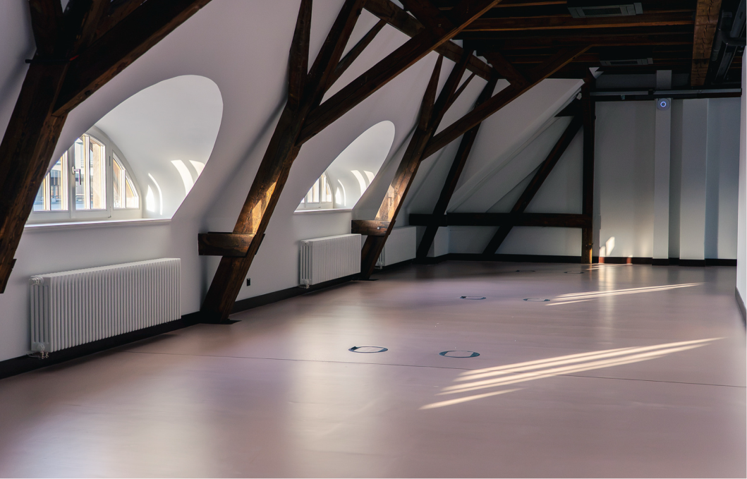 Leerer heller Raum mit schräger Decke, sichtbaren dunklen Holzbalken und drei großen halbrunden Fenstern mit Heizkörpern darunter, Sonnenlicht fällt auf den Boden.