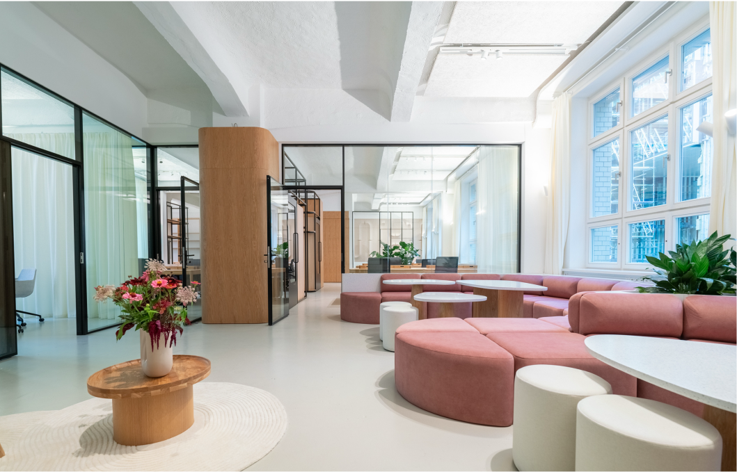 Helles, modernes Büro mit pinkfarbenen modularen Sitzgelegenheiten, runden weißen Hockern, mehreren Tischen und großen Fenstern. Links sind Glastüren zu weiteren Arbeitsräumen und ein runder Holztisch mit einem Blumenstrauß in einer Vase.