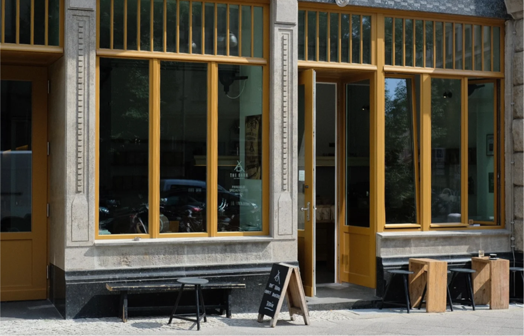 Außenansicht eines Cafés oder kleinen Geschäfts mit großen Fenstern und offenen gelben Türen, davor stehen zwei Holztische mit schwarzen Hockern und eine schwarze Tafel mit weißer Schrift auf dem Bürgersteig.