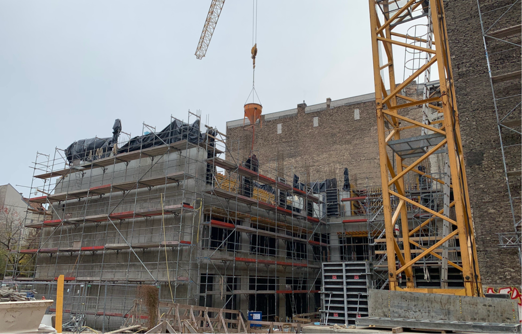 Baustelle eines mehrstöckigen Gebäudes mit Betonskelett und Stahlgerüst, ein gelber Baukran hebt eine orangefarbene Betonschütte, im Hintergrund eine alte Ziegelsteinmauer.