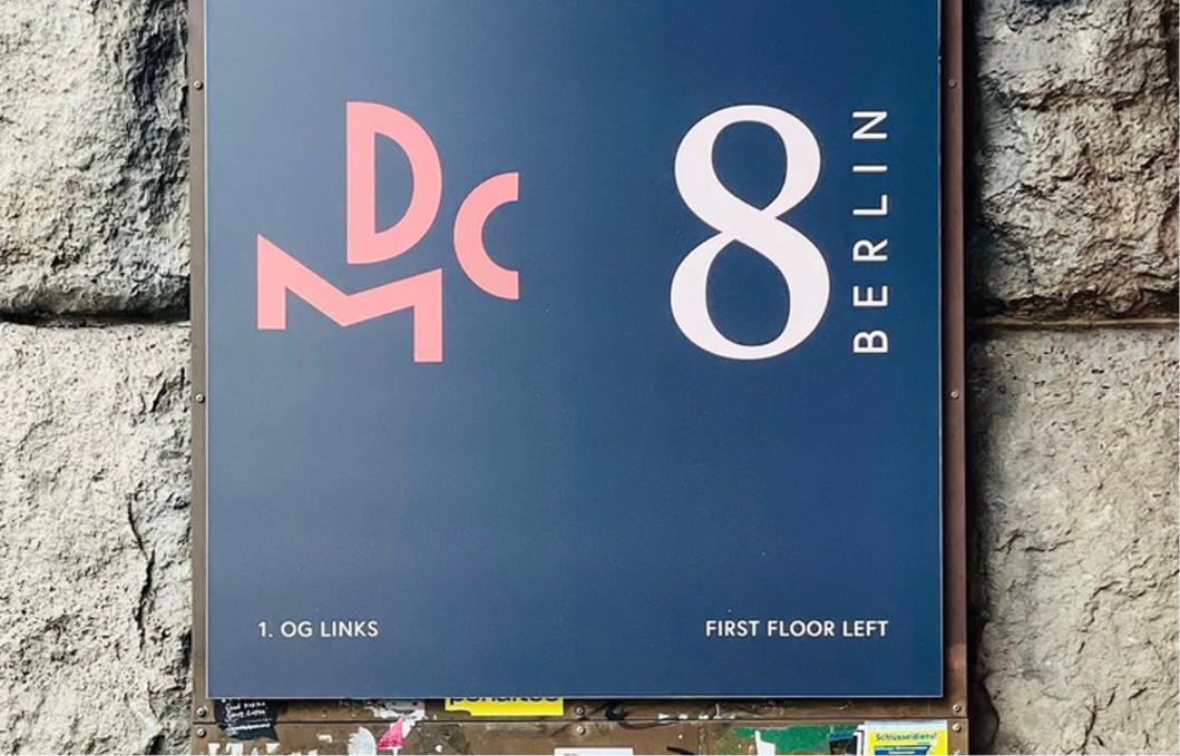 Schild an einer Steinmauer mit der Aufschrift 'DCM 8 BERLIN', darunter steht '1. OG LINKS' und 'FIRST FLOOR LEFT'.