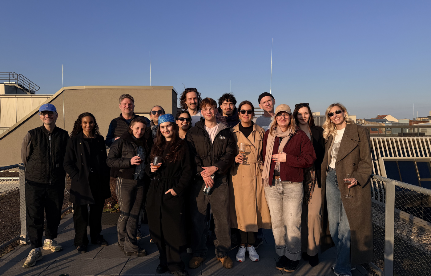Gruppenfoto mit fünfzehn Personen, die auf einer sonnenbeschienenen Dachterrasse stehen, in entspannter Atmosphäre mit lächelnden Gesichtern, einige halten Getränke in den Händen, klarer blauer Himmel im Hintergrund.