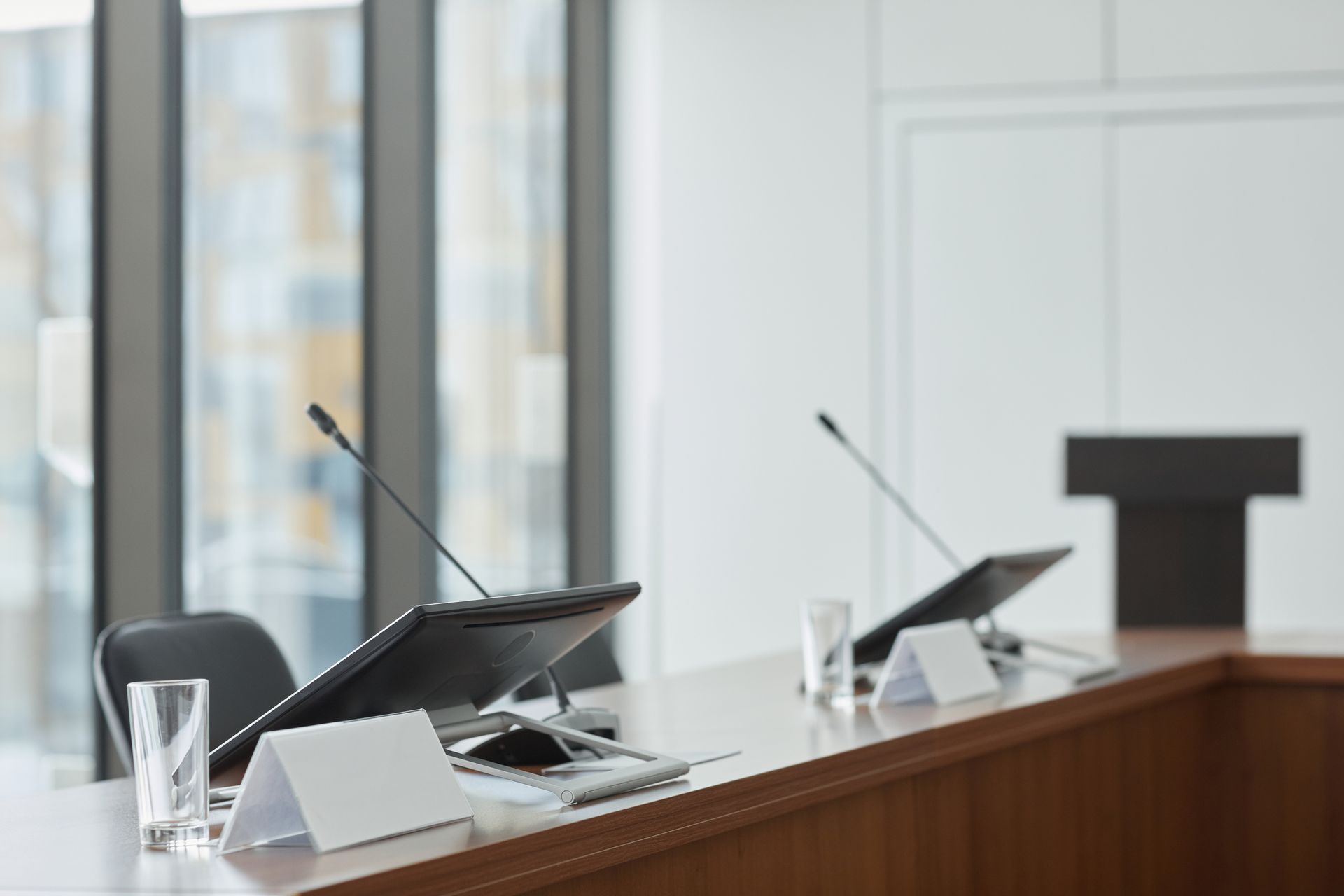 Empty conference table with microphones, screens, nameplates, and glasses near large windows.