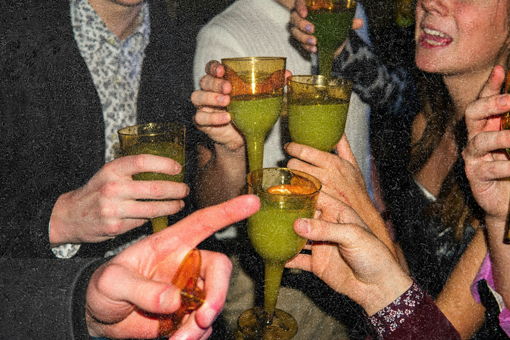 Group of people clinking yellow stemmed glasses filled with a green drink in celebration.