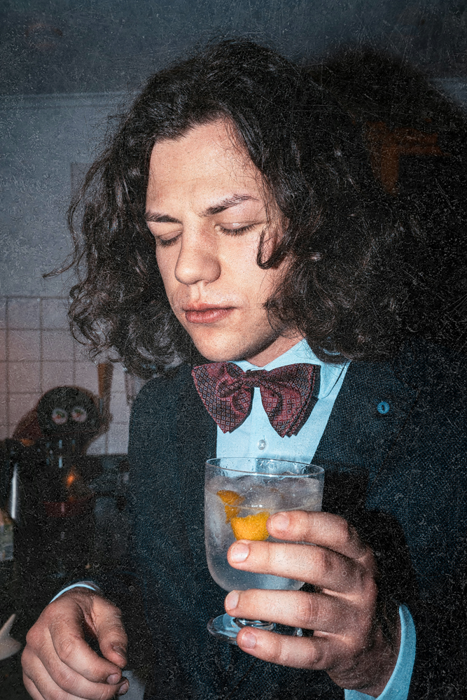 Young man with long hair and bow tie holding a glass of iced beverage, eyes closed.