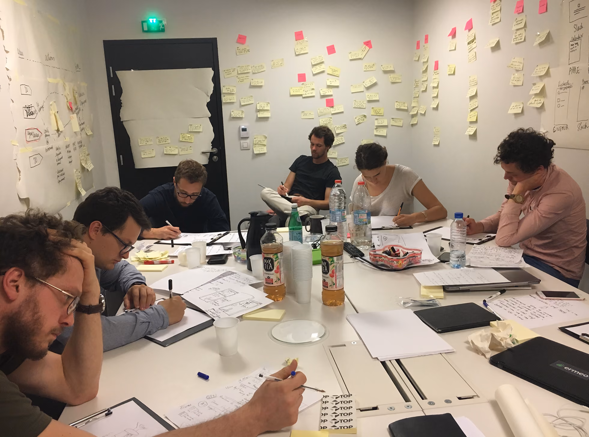 Participants assis autour d'une grande table en train de dessiner, murs couverts de post-it