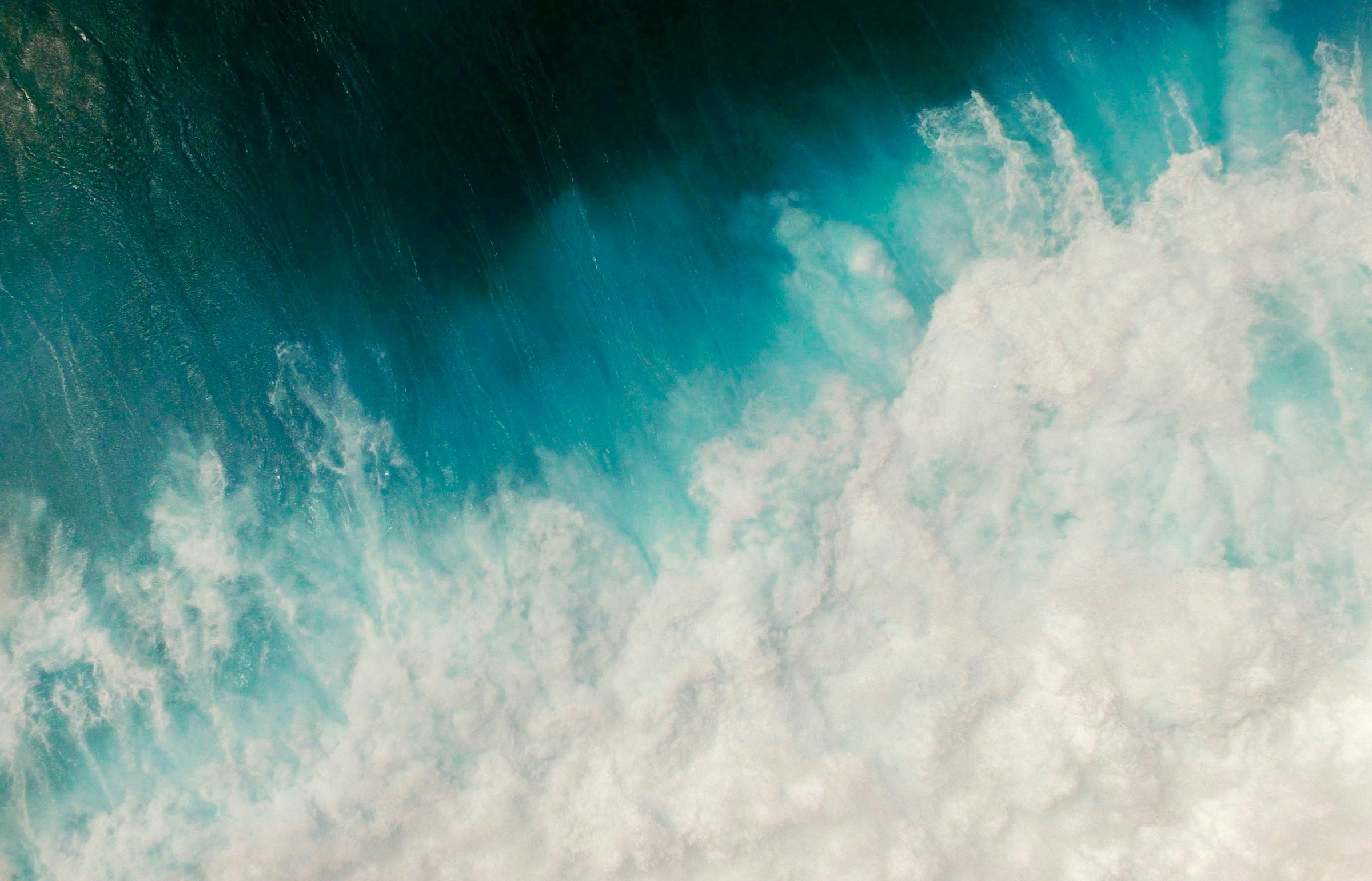 Aerial view of a breaking ocean wave, deep turquoise water meeting white foam