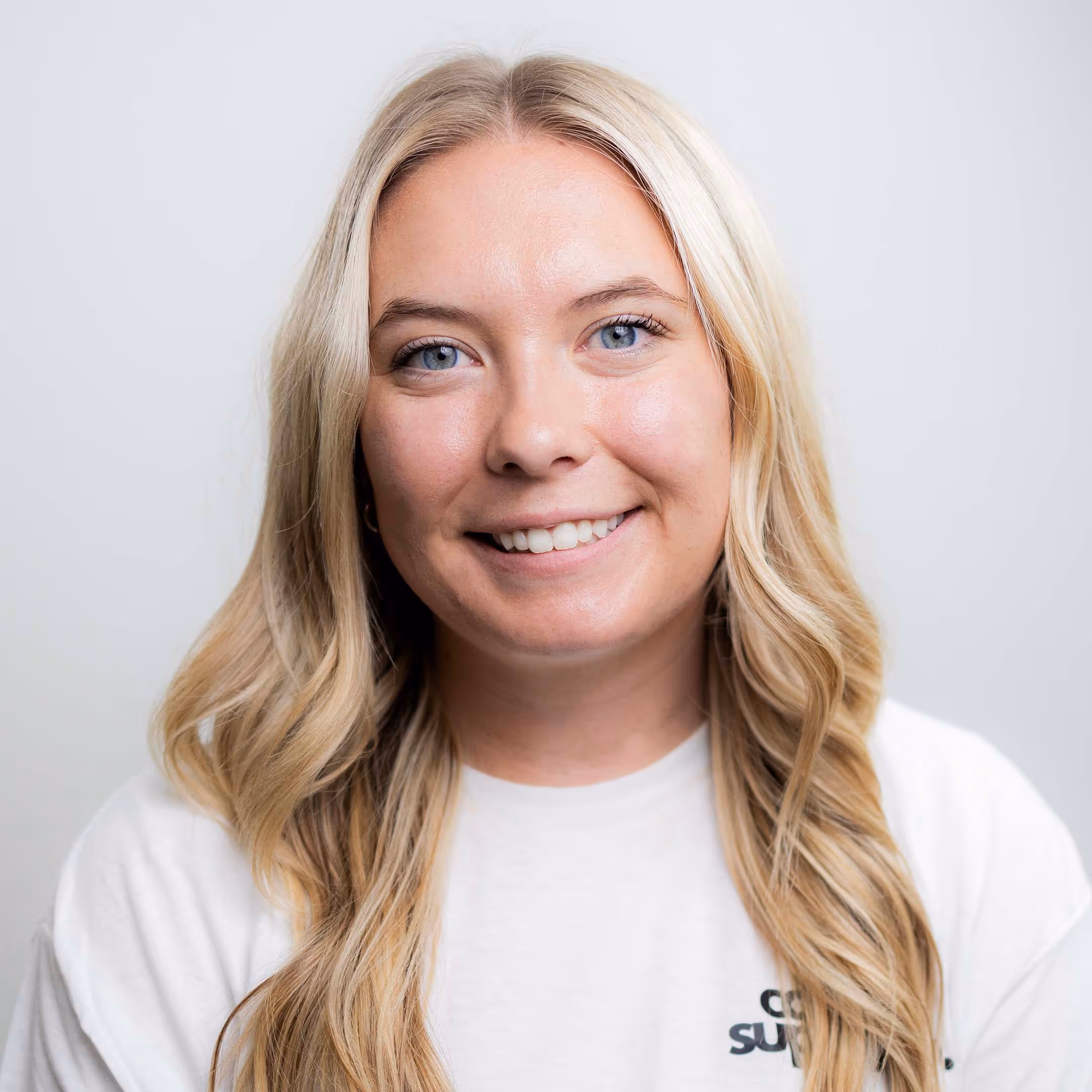 Smiling young woman with long blonde hair wearing a white shirt against a plain light background.