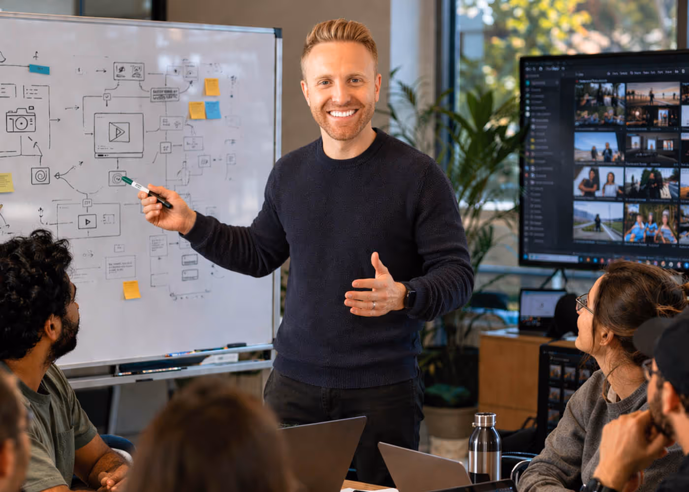 Man explaining a workflow diagram on a whiteboard to a group seated around a table with laptops in a modern office.