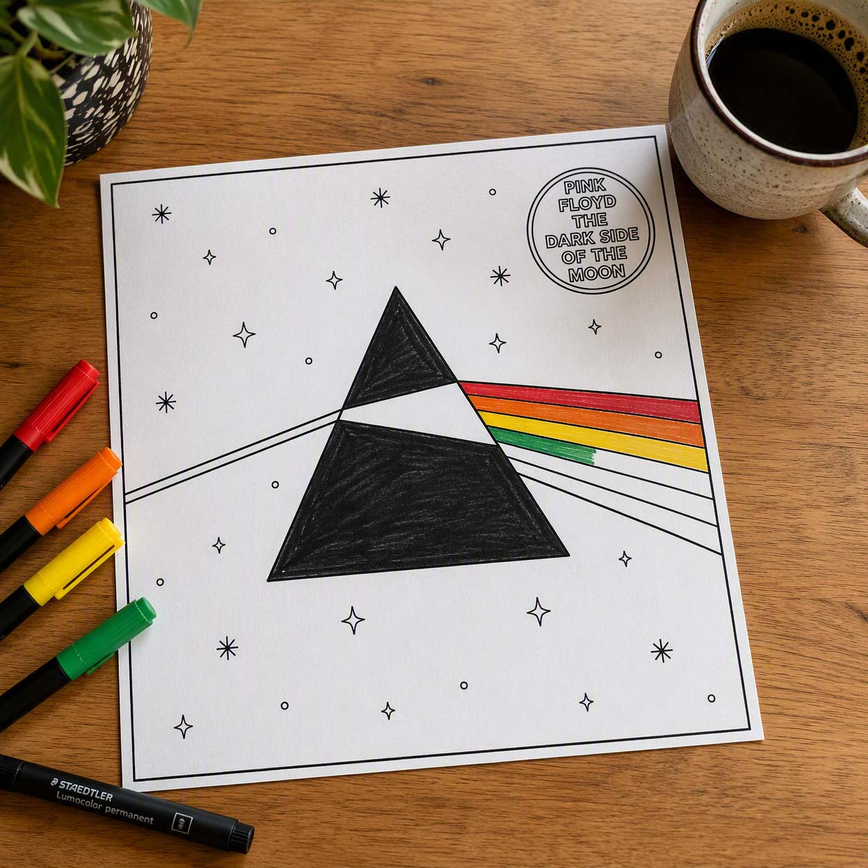 A colouring book version of Dark Side of the Moon Album cover on a desk with pens and a coffee.