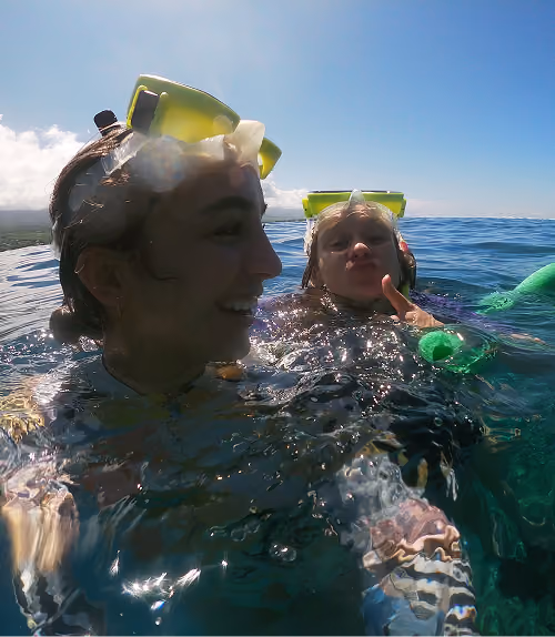 Two people in snorkel gear smiling and floating in clear ocean water under a blue sky.