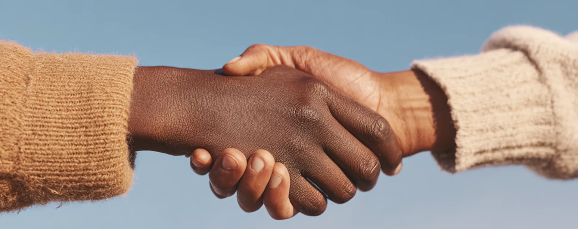 Two people shaking hands, one with dark skin and the other with light skin, both wearing sweaters against a blue sky.