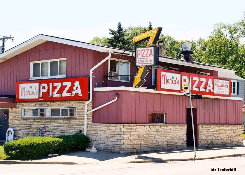 Corner building with red siding and stone base displaying multiple signs for Maria's Pizza and Italian foods.