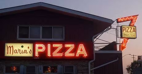 Neon sign reading 'Maria's PIZZA' and arrow-shaped sign that says 'PIZZA Orders To Go' on a building at dusk.