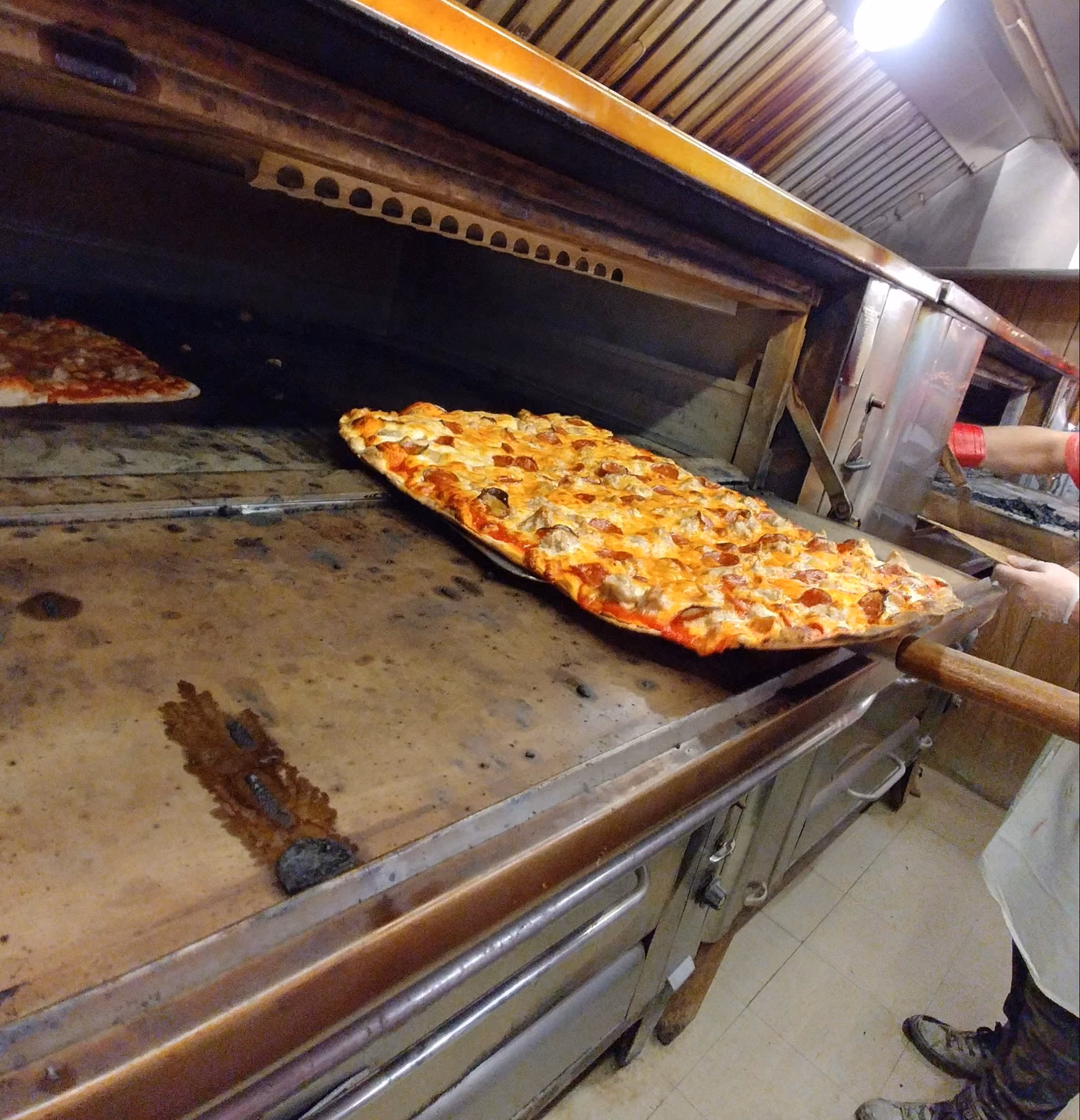 Person using a wooden peel to place a rectangular pizza with cheese and sausage into a pizza oven.