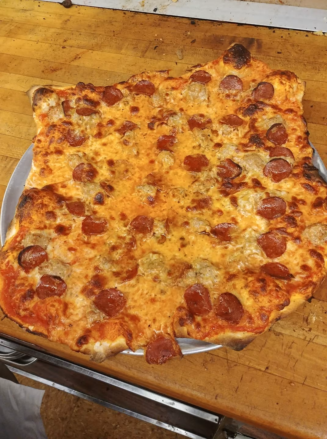 Large pepperoni and sausage pizza with uneven crust on a metal tray on a wooden surface.