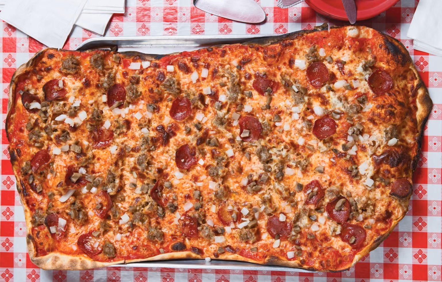 Large rectangular pepperoni, sausage, and onion pizza on a red and white checkered tablecloth.