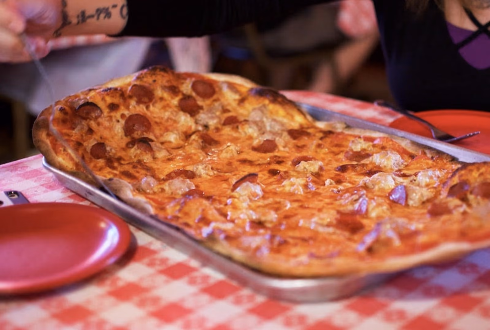 Large pepperoni and sausage pizza on a silver tray on a red and white checkered tablecloth.