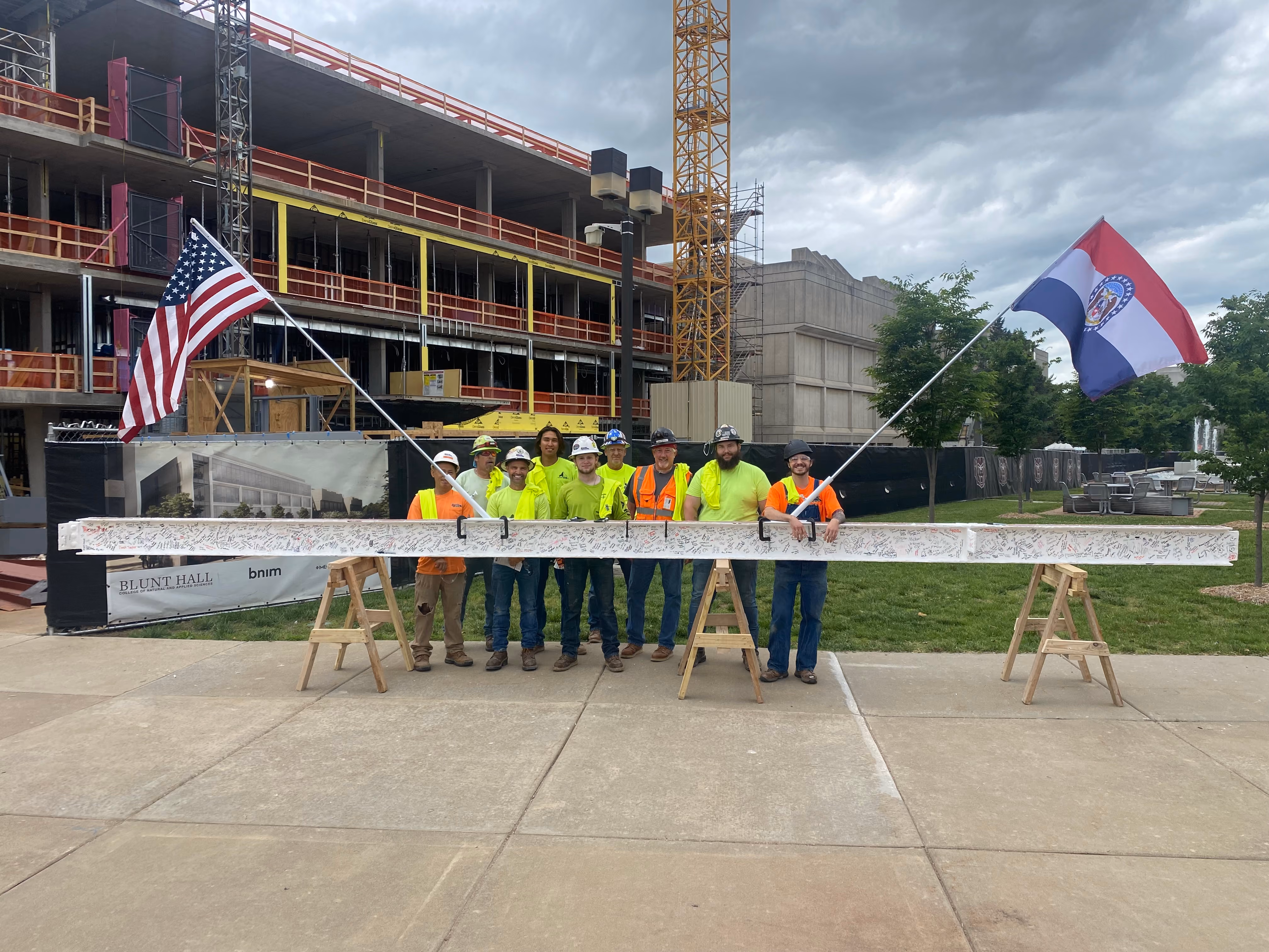 Group of workers in high-visibility gear posing with a flag at a building site