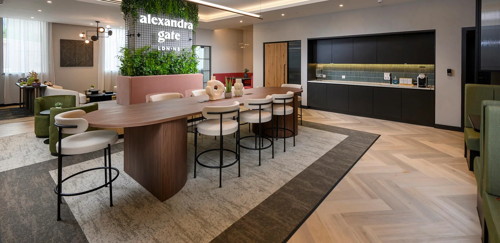 Modern lounge with oval table, cream chairs, green plants, and black kitchen cabinets under warm lights.