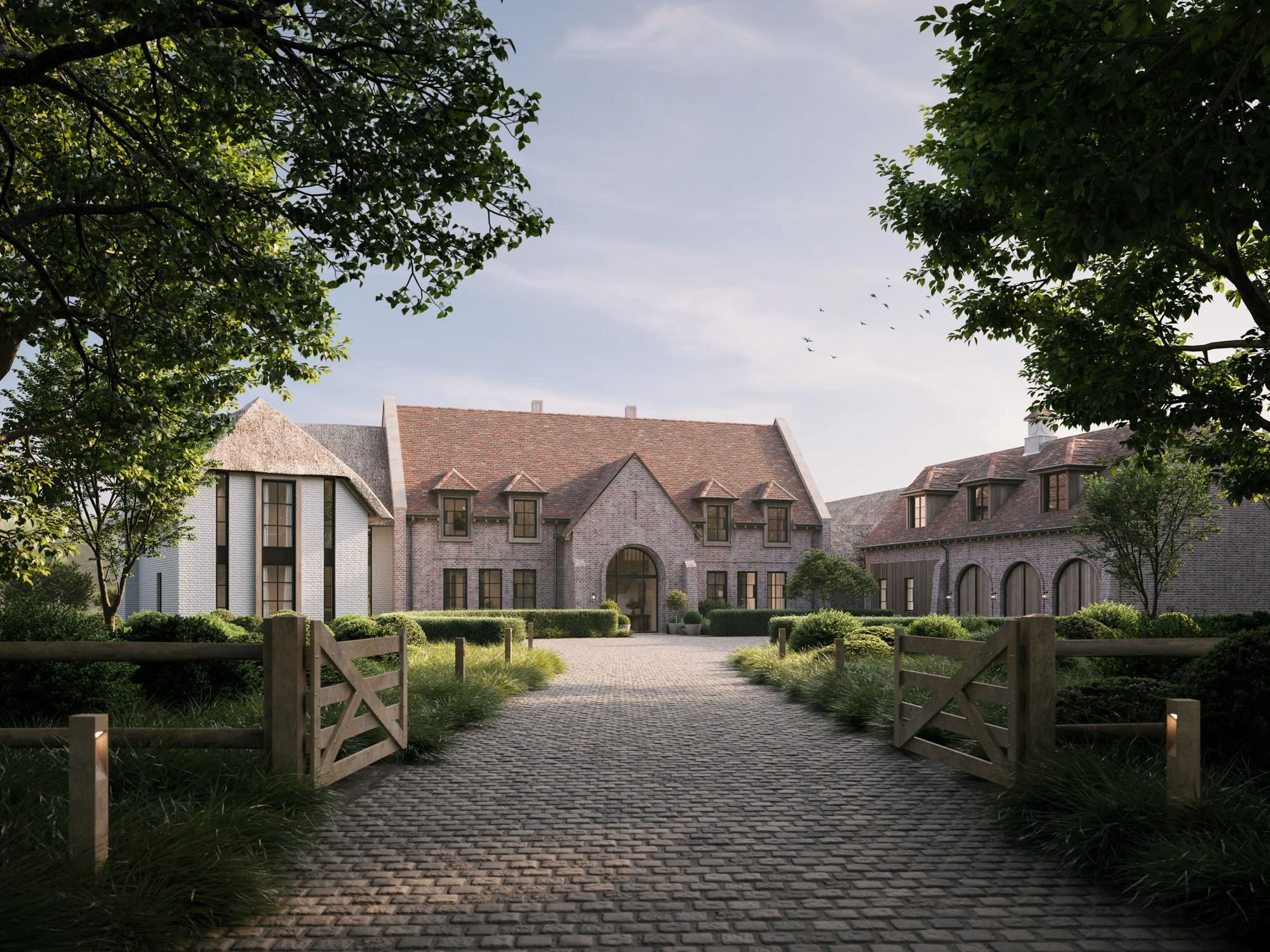 A large, elegant countryside house with brick and white plaster walls, red tile roofs, multiple windows, and a central arched entrance, framed by green hedges and trees, with a cobblestone driveway and wooden gates in the foreground under a partly cloudy sky.