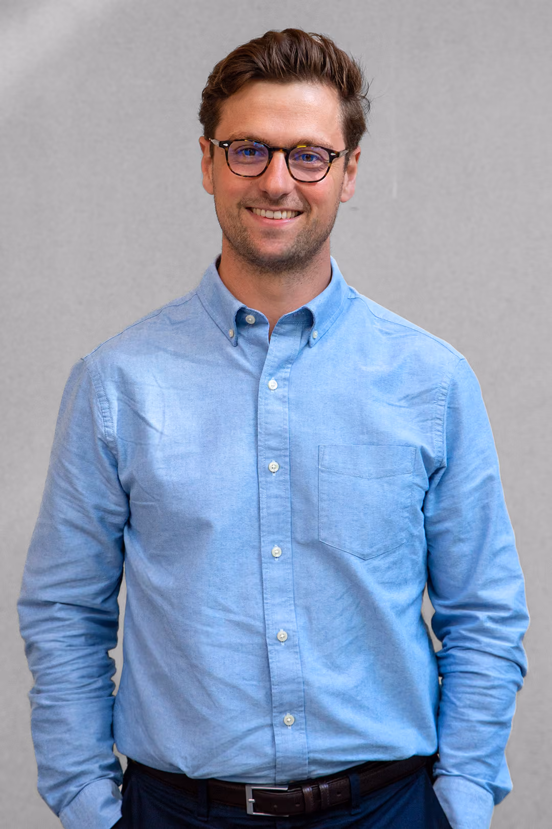Smiling man with glasses wearing a light blue button-up shirt against a plain gray background.