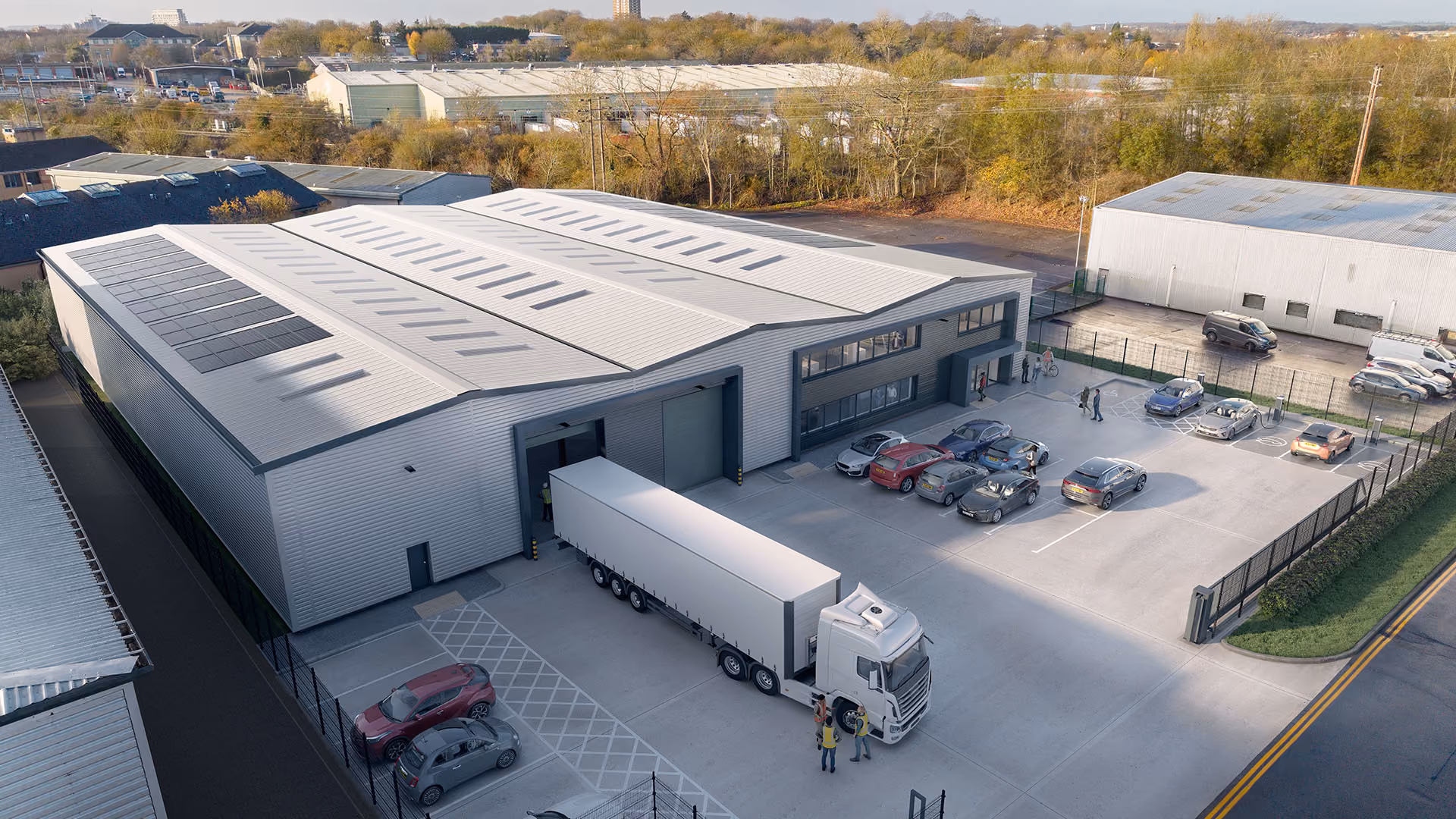 Warehouse with solar panels on roof, a large truck docked, cars in parking, and people near entrance.