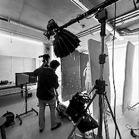 A black and white behind-the-scenes photo of e-commerce photography at LRP studios, showing a camera operator filming a model posing between tall reflectors under an overhead studio light.